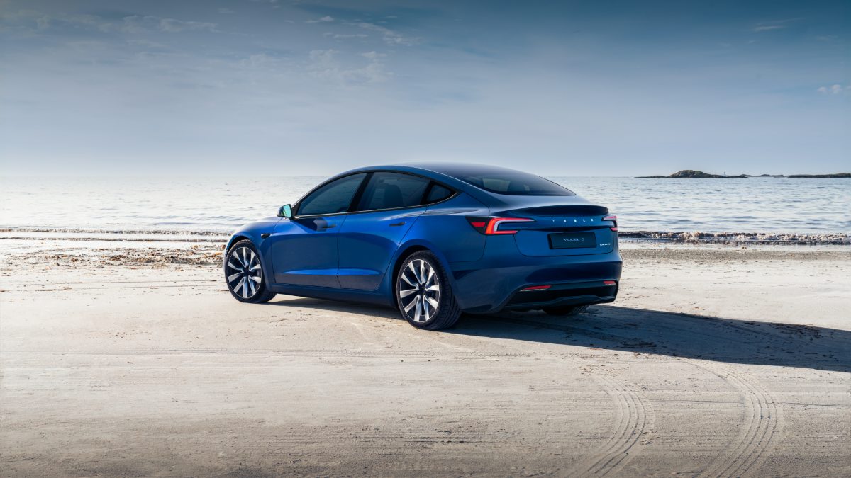 Electric blue Tesla Model 3 sedan photographed from rear three-quarter angle on sandy beach, featuring sleek aerodynamic design and distinctive LED taillights.