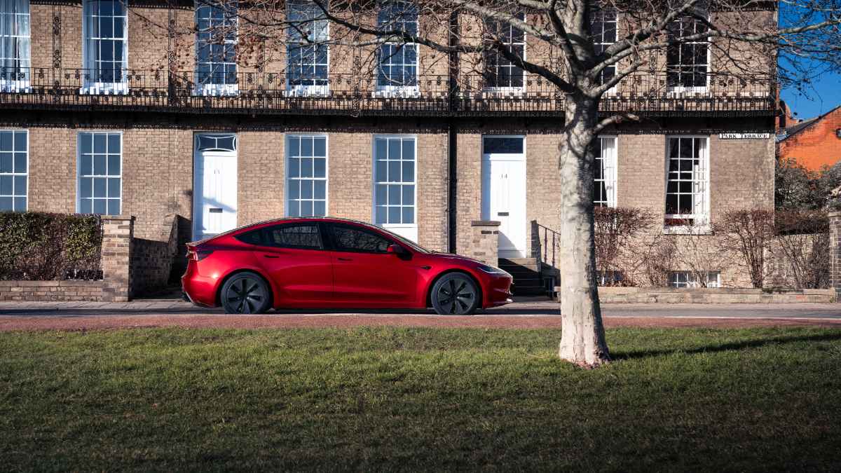 A red Tesla Model 3 is shown from the side profile, parked on a street in front of a classic brick townhouse building with white-trimmed windows.