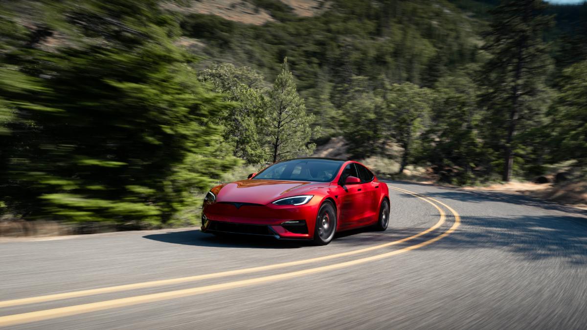 Red 2026 Tesla Model S driving on a winding mountain road surrounded by trees and greenery