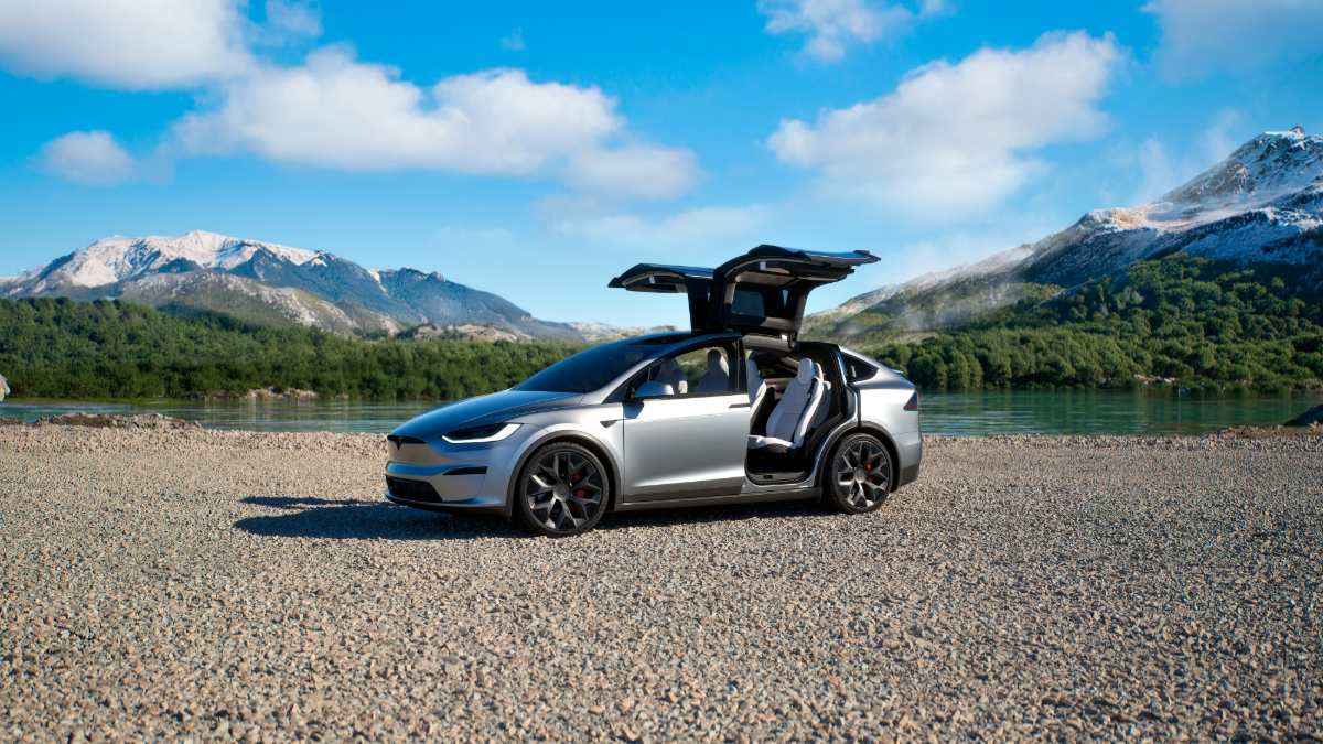 A silver Tesla Model X with its distinctive falcon-wing doors open, photographed from a three-quarter front angle against a stunning backdrop of snow-capped mountains and a turquoise lake.