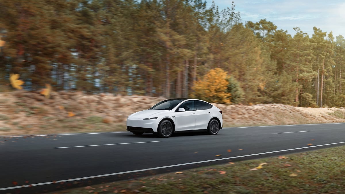 A white 2026 Tesla Model Y speeds down a forest road. The background is filled with tall trees and autumn leaves, creating a dynamic and serene atmosphere.