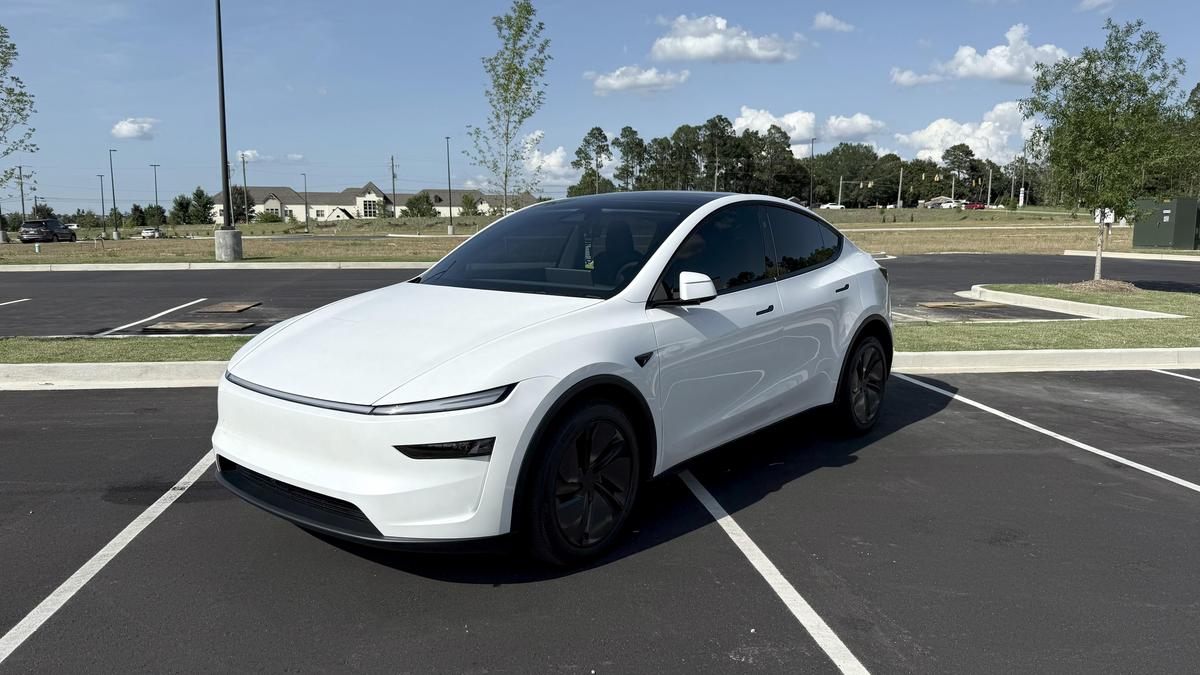 White Tesla Model Y front three-quarter view parked in an empty parking lot on a sunny day.