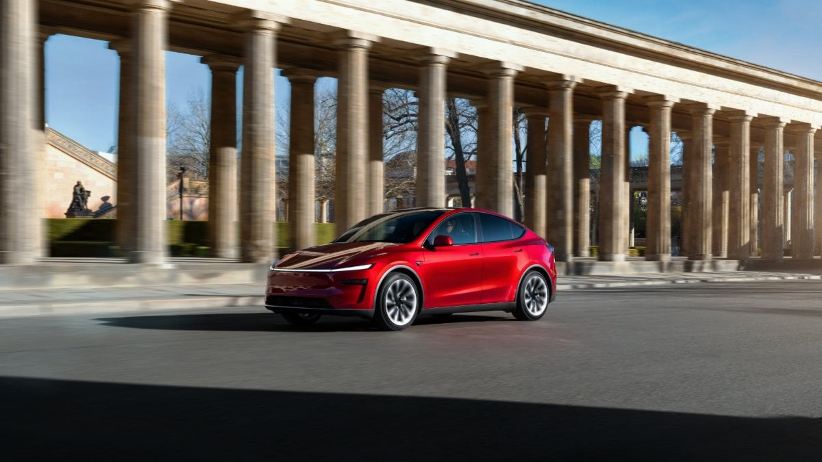 Red electric car driving past historic stone columns on a sunny day.