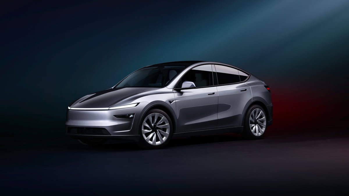 Photograph of a 2026 Tesla Model Y electric vehicle in a Juniper silver color, captured from the front three-quarter angle against a colorful background.