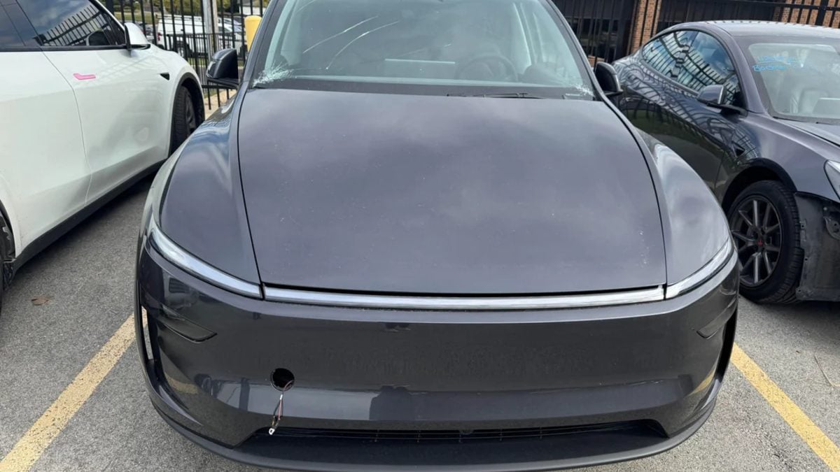 2026 Tesla Model Y Juniper in metallic gray, front quarter view, showing distinctive curved hood and chrome trim strip, parked in lot beside white vehicle.
