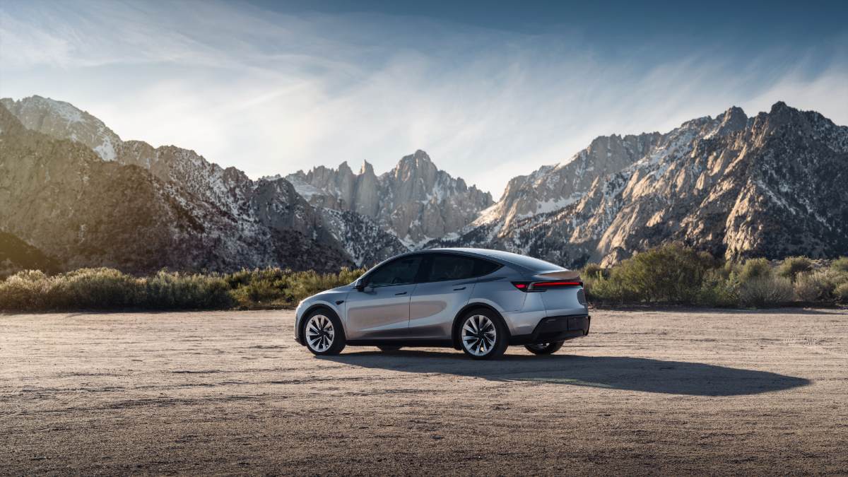 Silver 2026 Tesla Model Y parked on desert ground with dramatic snow-capped mountain range in background, rear three-quarter view