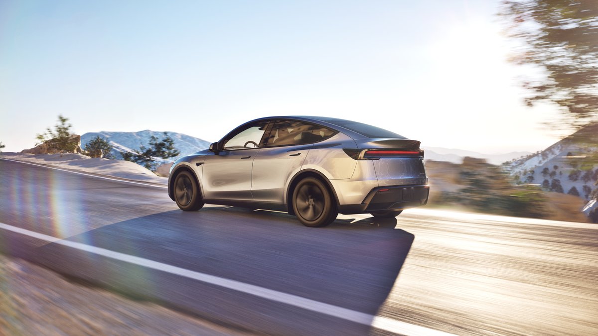Photograph of a 2026 Tesla Model Y electric SUV in the Juniper color from a 3/4 rear angle, showing the vehicle's silver exterior color, rear lights, and overall shape and design.