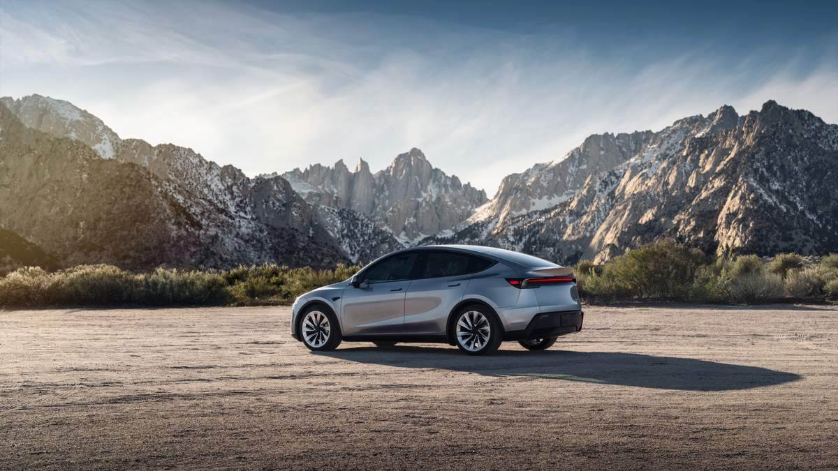 2026 Tesla Model Y Juniper in silver, rear three-quarter view parked on open terrain with dramatic mountain landscape.