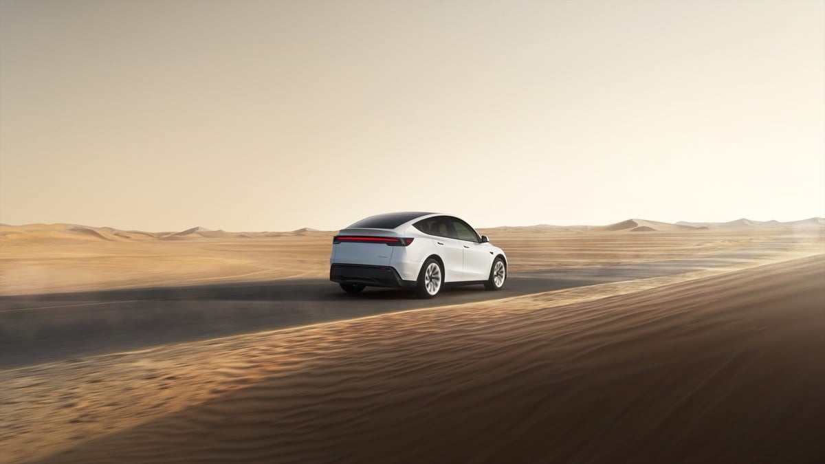 2026 Tesla Model Y Juniper in white, rear three-quarter view driving through desert landscape with sand dunes at sunset.