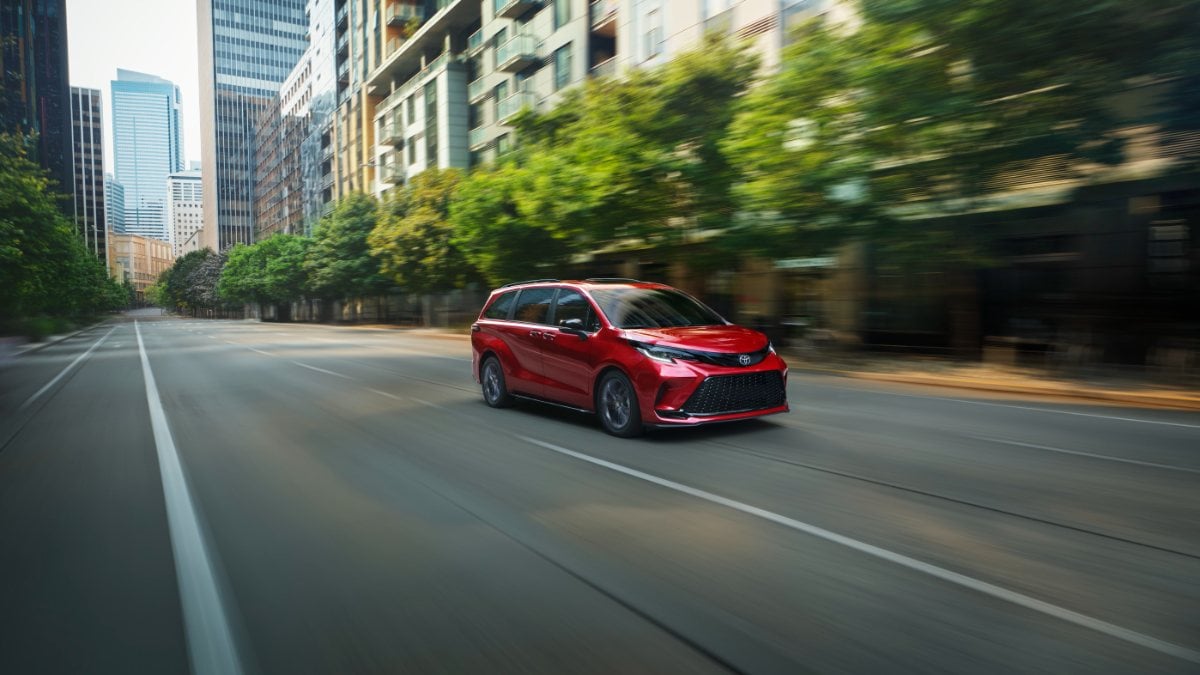 A red 2024 Toyota Sienna minivan shown from the front three-quarter angle driving on a city street with motion blur, surrounded by urban buildings and trees.