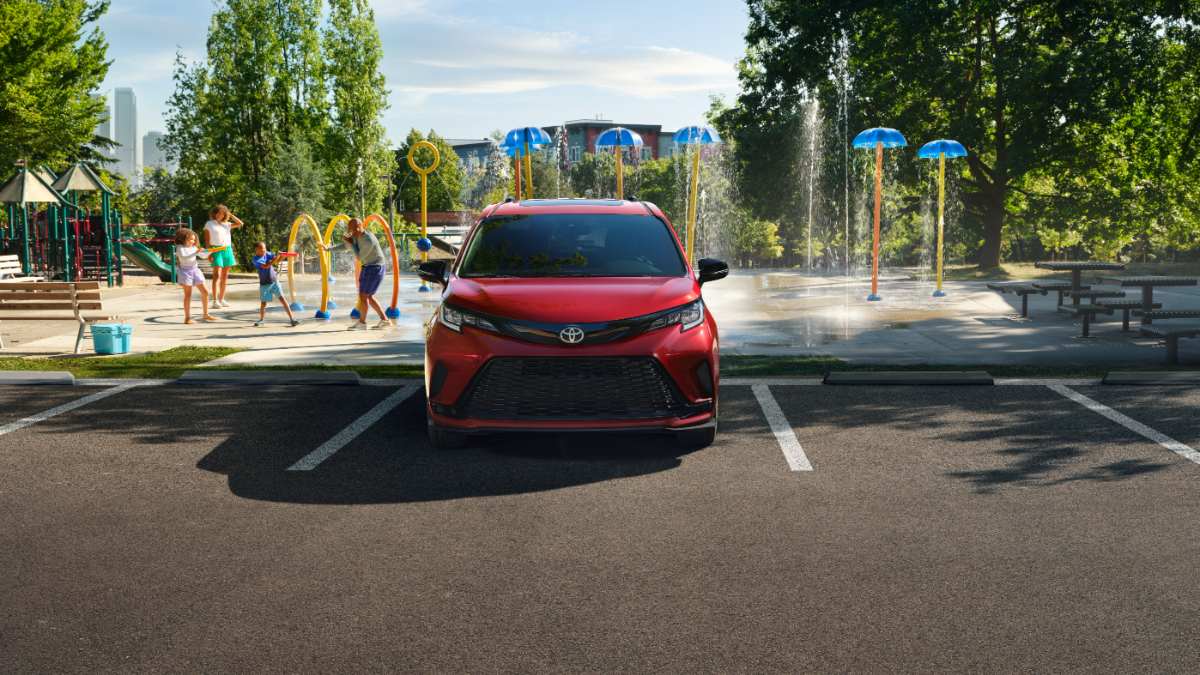 A red 2023 Toyota Sienna minivan is shown from the front quarter view, parked in a lot near a splash pad playground with water features and city buildings visible in the background.