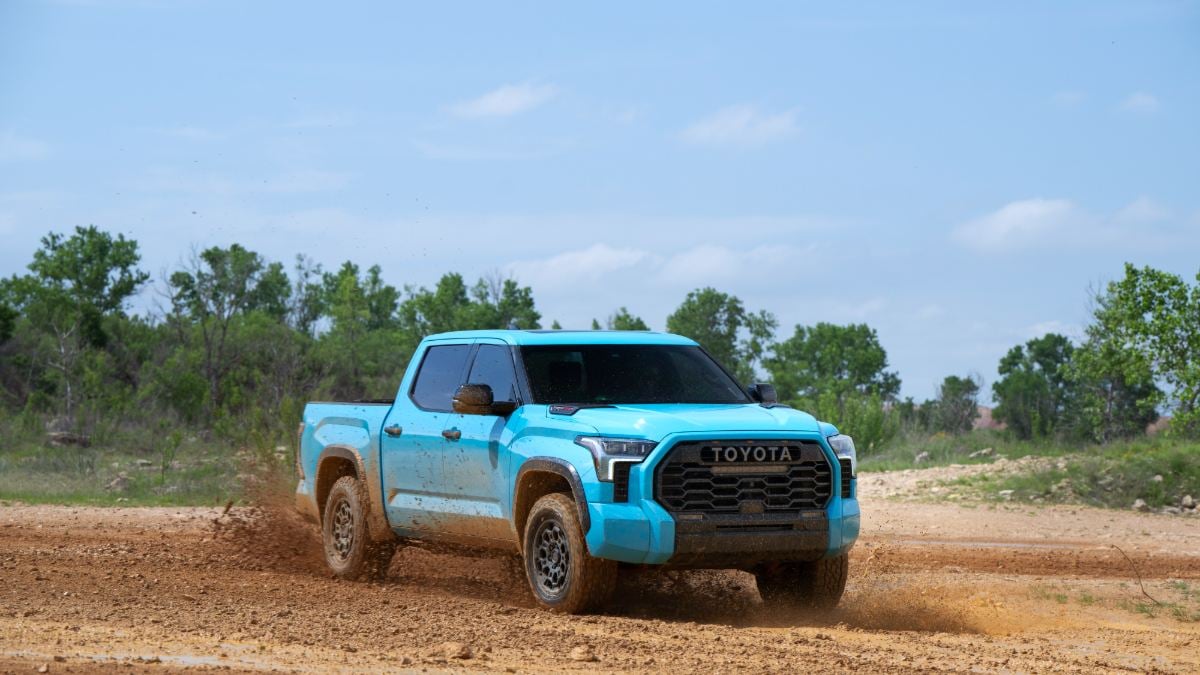 A bright blue 2024 Toyota Tundra shown from a front three-quarter angle, kicking up dirt while off-roading on a muddy trail with trees in the background.