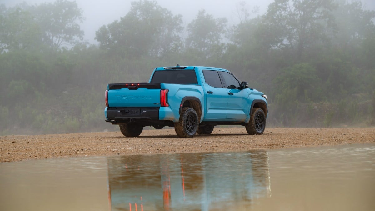 A bright turquoise 2026 Toyota Tundra TRD Pro pickup truck is shown from a rear three-quarter angle, parked on dirt terrain with its reflection visible in a puddle of water on a misty day.