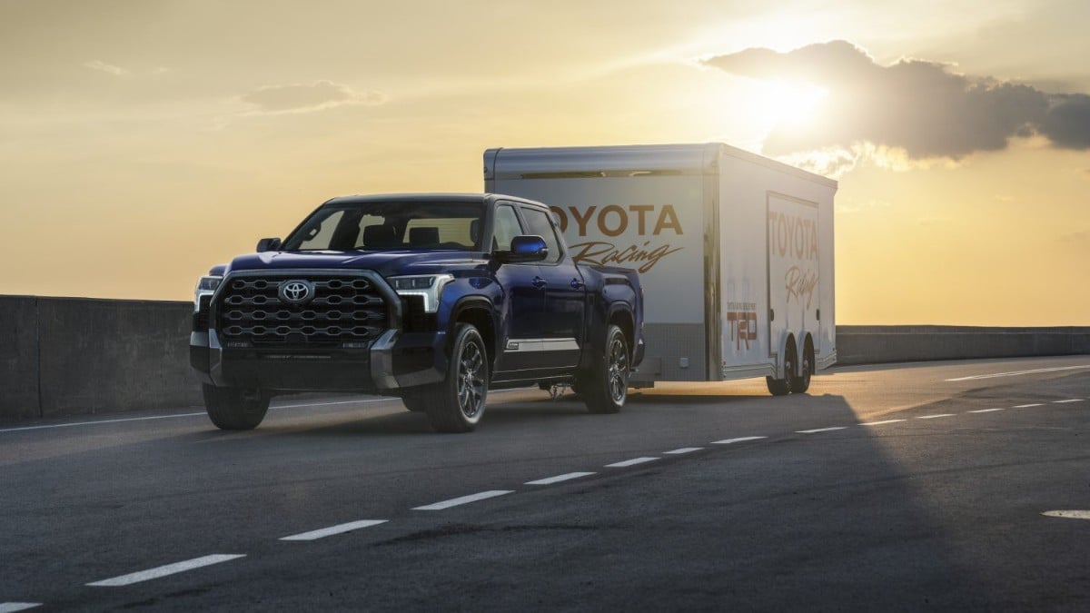 A dark blue 2024 Toyota Tundra pickup truck is shown from a front three-quarter angle at sunset, parked near Toyota Racing transport trailers with dramatic golden sky in the background.