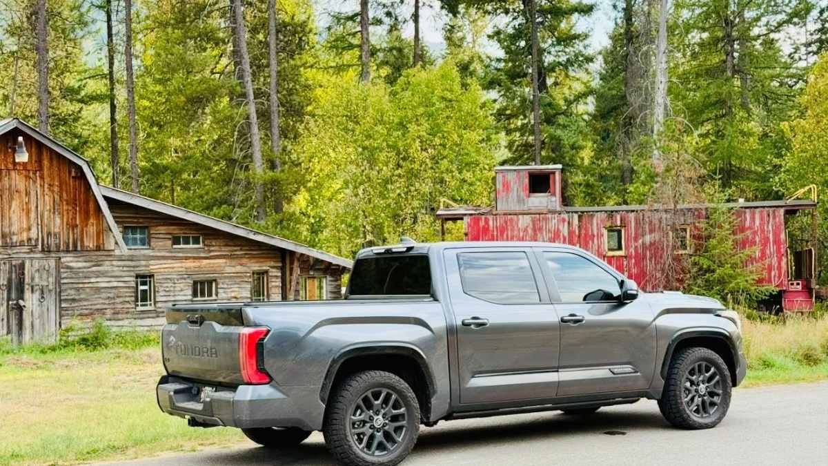 A gray Toyota Tundra pickup truck, likely a 2022 or 2023 model, is shown from the rear quarter view parked in front of a rustic wooden cabin with red accents surrounded by evergreen forest.