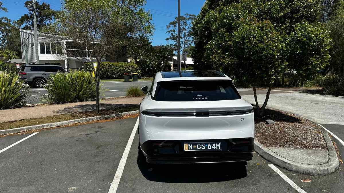 White car parked in a lot, surrounded by trees and a residential street.