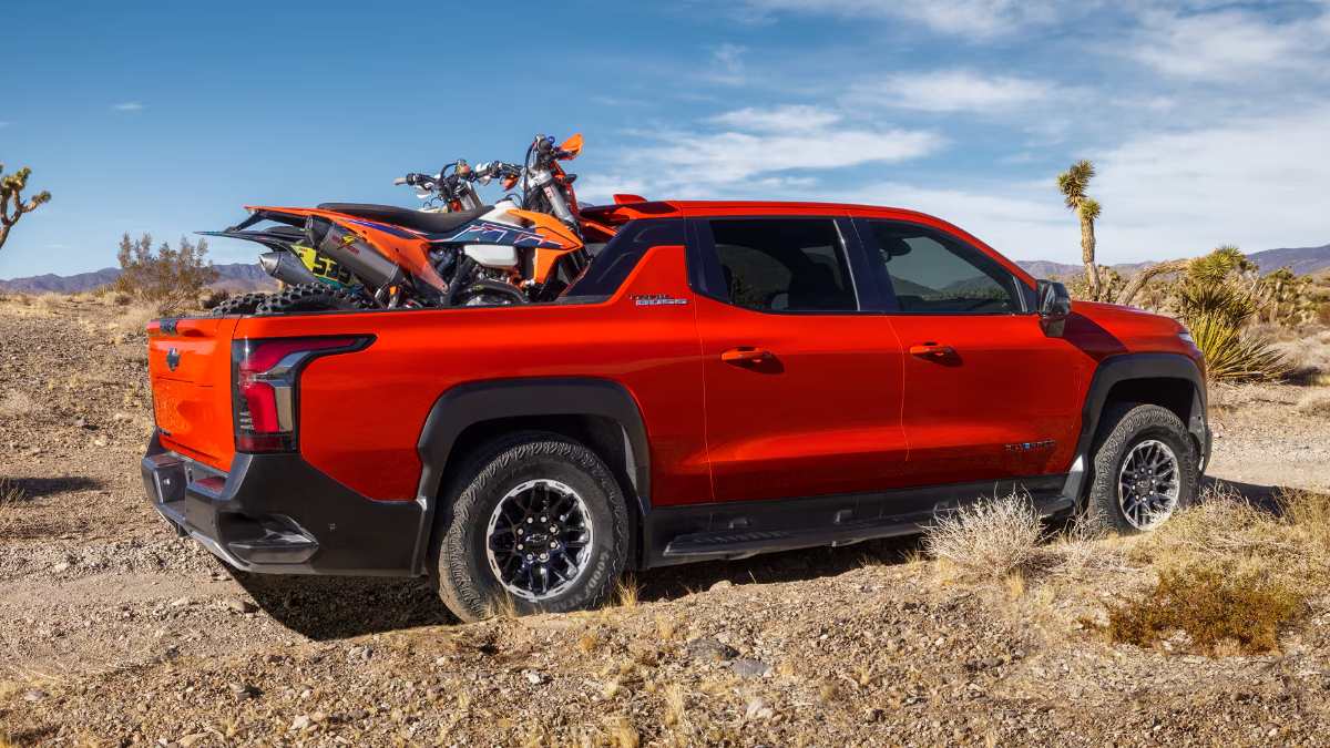 2026 Chevrolet Silverado EV red electric truck rear three-quarter view parked in desert with dirt bikes loaded in the bed.