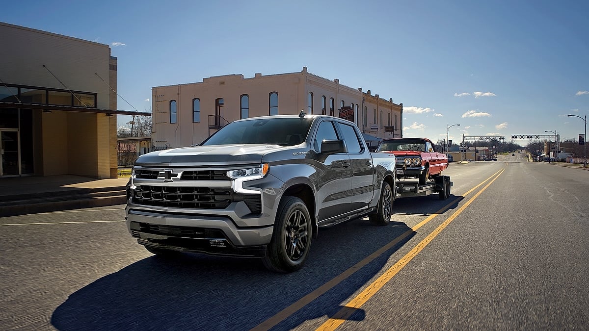 2026 Chevrolet Silverado towing a classic red truck on a flatbed trailer through a city street.