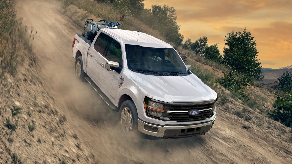White 2026 Ford F-150 driving along a dusty mountain trail at sunset.