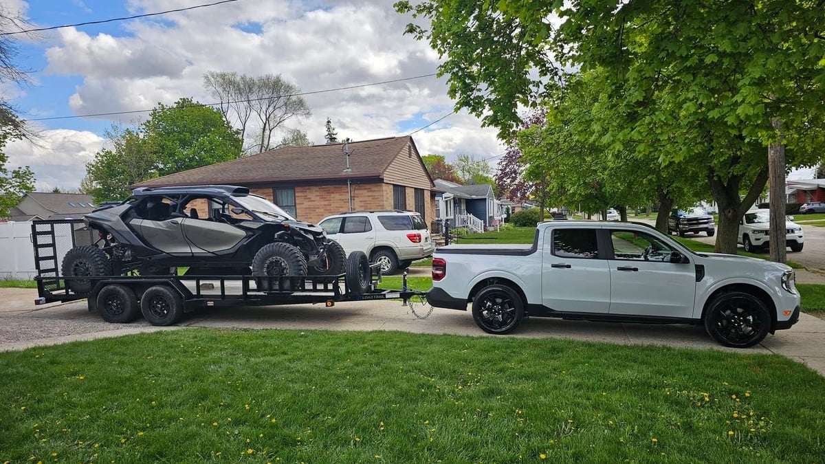 2026 Ford Maverick Hybrid towing an ATV parked neatly in front of a modern suburban neighborhood driveway