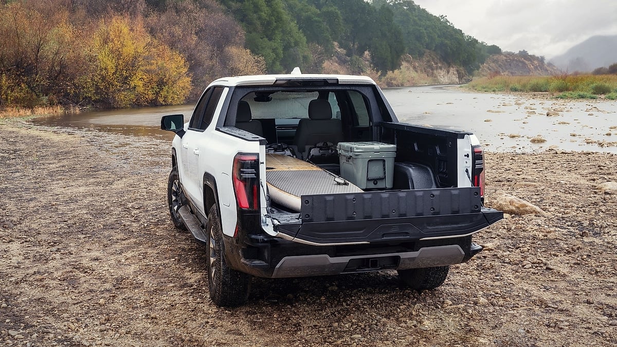 White 2026 GMC Sierra EV AT4 parked near a rocky riverbed with the tailgate and MidGate open and camping gear in the bed.