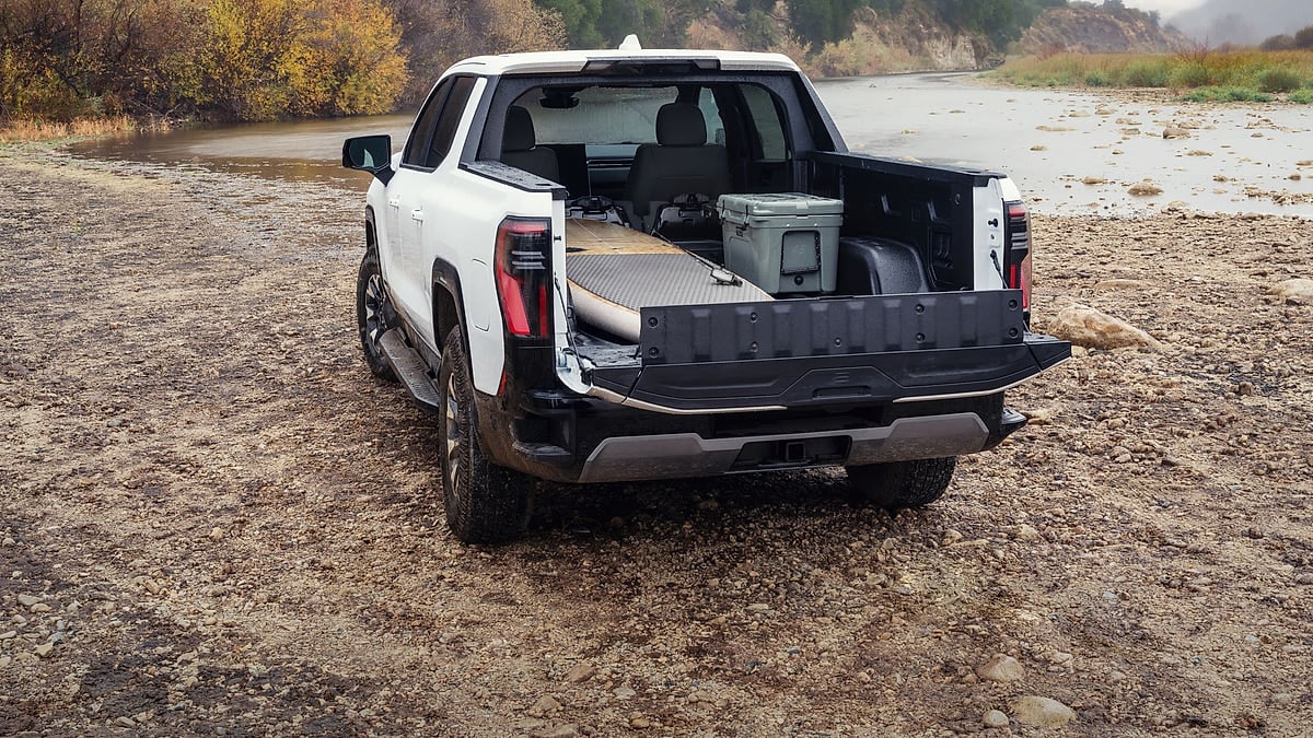 2026 GMC Sierra EV AT4 parked on a rocky riverbed with its tailgate and MidGate open, showing cargo in the truck bed.