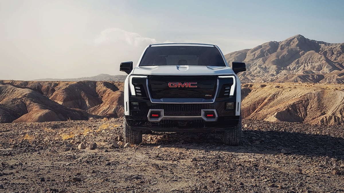 White 2026 GMC Sierra EV AT4 shown from the front on rocky desert terrain with mountains in the background.