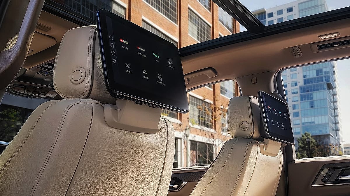 Interior view of a 2026 GMC Yukon showing rear-seat entertainment screens mounted behind the front headrests, with a panoramic sunroof and premium light-colored leather seating visible.
