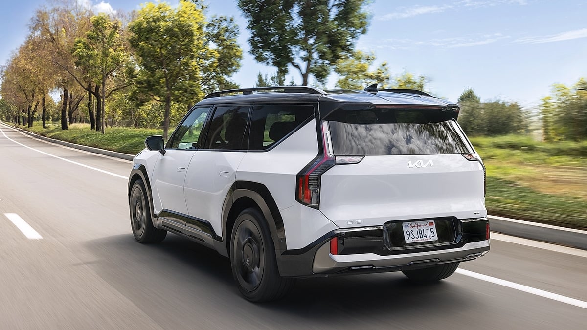2026 Kia EV9 electric SUV in white driving on a tree-lined road, rear three-quarter view