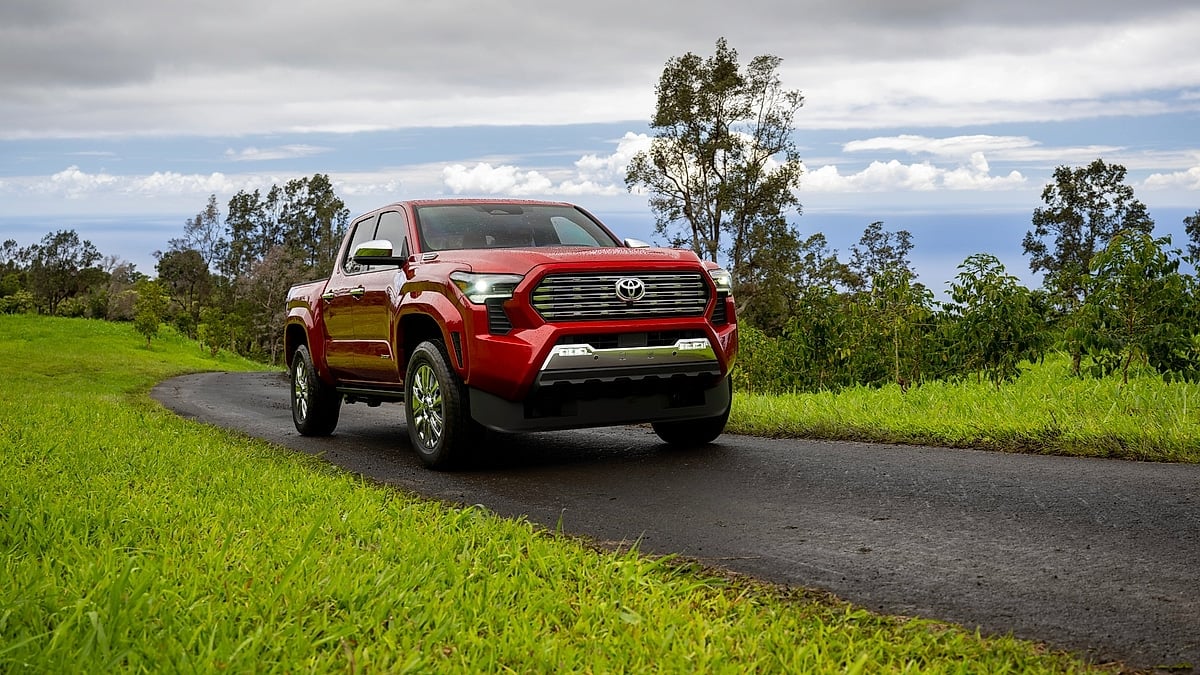 Red 2026 Toyota Tacoma Limited driving on a narrow road with trees, ocean and cloudy sky in the background.