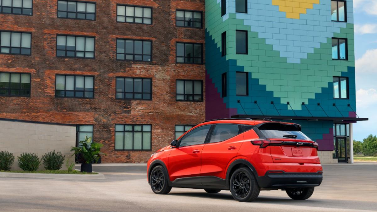 2027 Chevrolet Bolt EV red compact electric SUV rear three-quarter view parked in modern urban setting with brick and colorful building backdrop