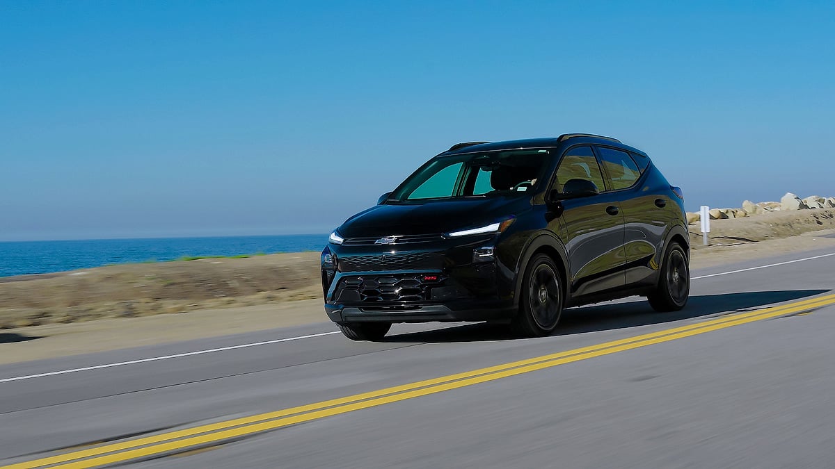 Black 2027 Chevrolet Bolt RS driving on a coastal road with the ocean in the background.
