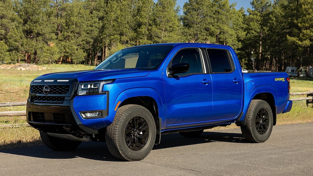 Blue 2027 Nissan Frontier Sport pickup parked on a paved road near a pine forest.