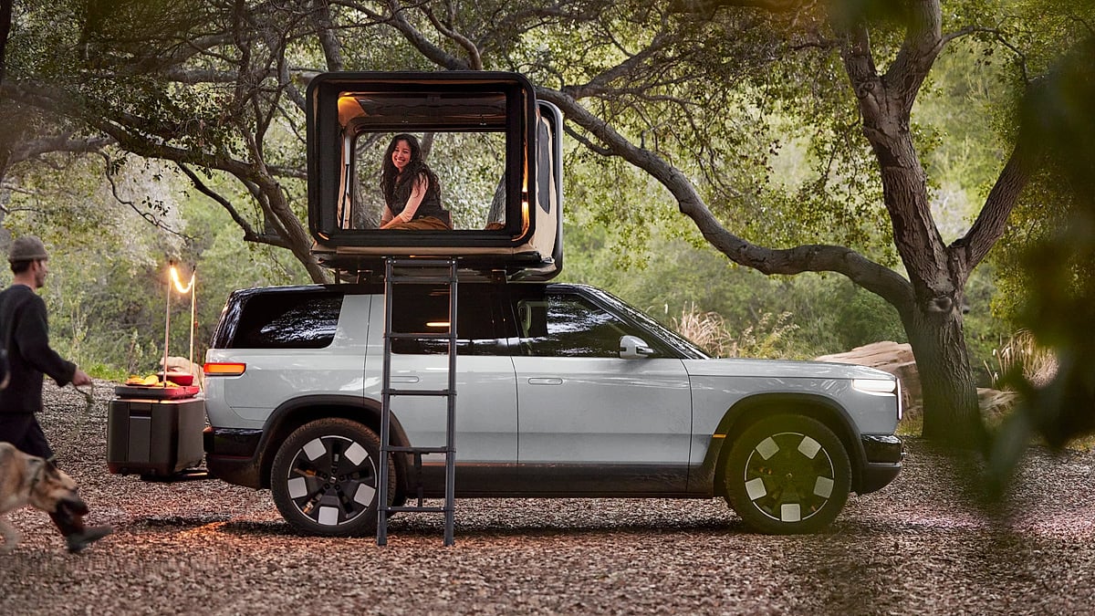 2027 Rivian R2 parked at a forest campsite with a rooftop tent open above the SUV.