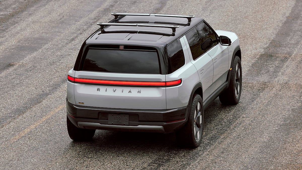 White 2027 Rivian R2 shown from a high rear three-quarter angle with roof crossbars and full-width taillight.