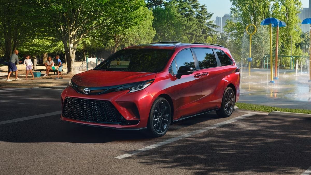 A red 2026 Toyota Sienna minivan shown from the front three-quarter angle, parked near a splash pad park with water features visible in the background.