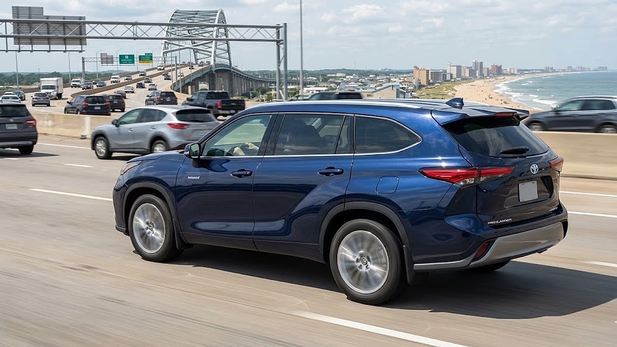 A dark blue 2026 Toyota Highlander Hybrid drives on a multi-lane highway in Virginia Beach, Virginia, with the Atlantic ocean and coastline in the background, without any text