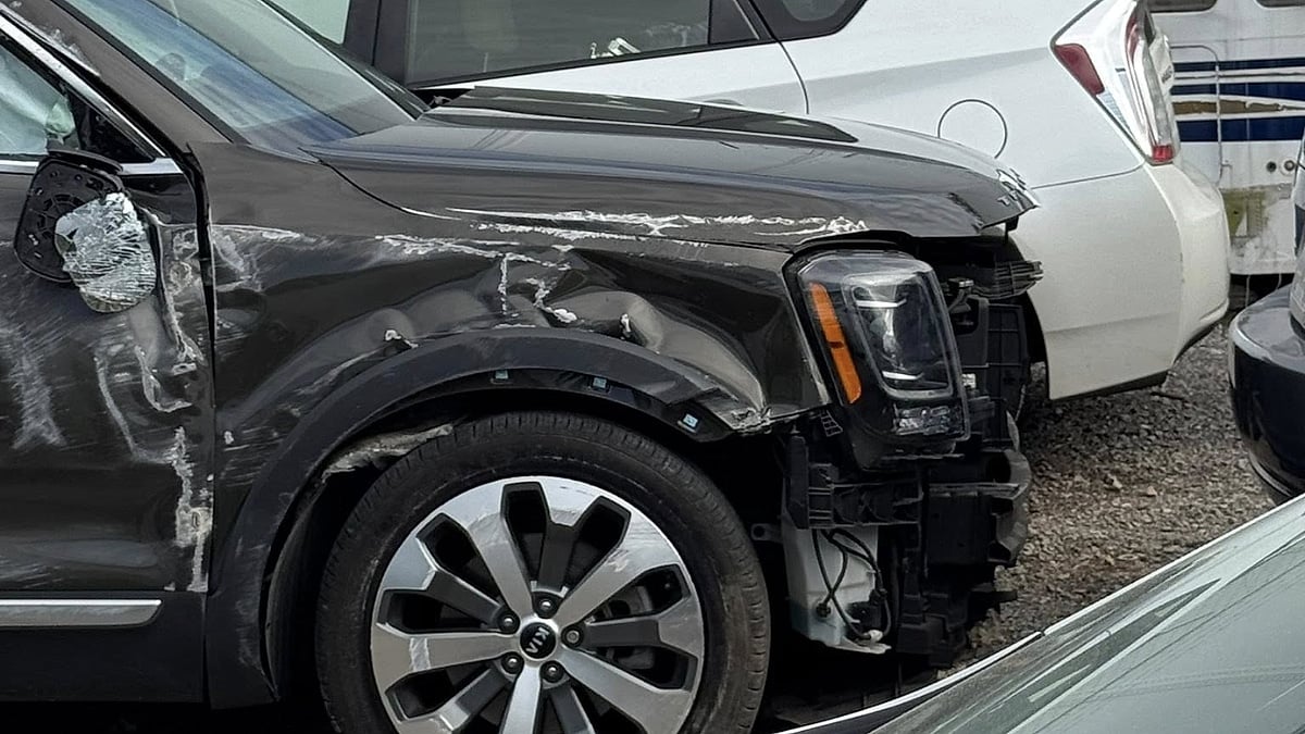 Jenni's 2020 Kia Telluride with a missing front bumper and side mirror, showing scratches and dents. Parked closely behind is a white vehicle. The scene conveys accident aftermath.