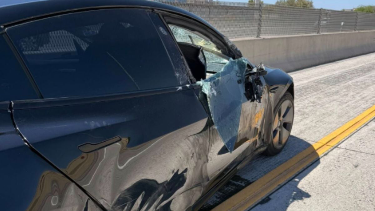 A black Tesla Model 3 with severe damage to the driver's side, featuring a shattered window and dented body, parked on a highway
