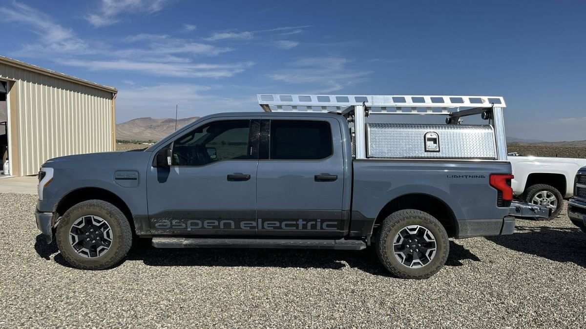A gray Ford F-150 Lightning with a utility rack is parked on gravel next to a beige building. The truck's side reads &quot;Aspen Electric.&quot; The sky is clear.