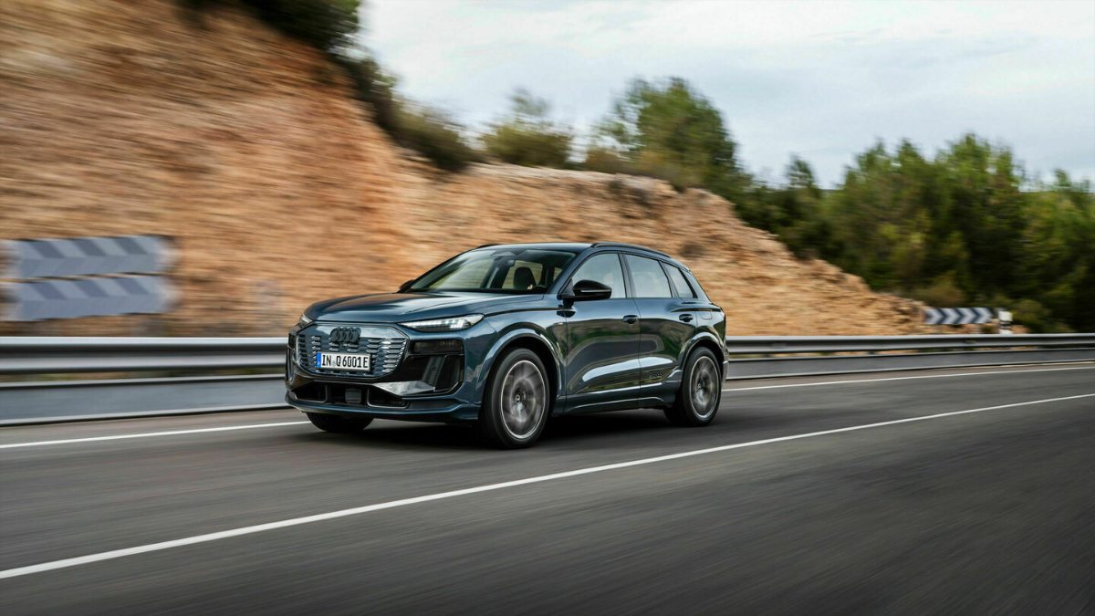 Audi Q6 e-tron in motion, front three-quarter view, dark color, showcasing distinctive grille and headlights on a winding road.