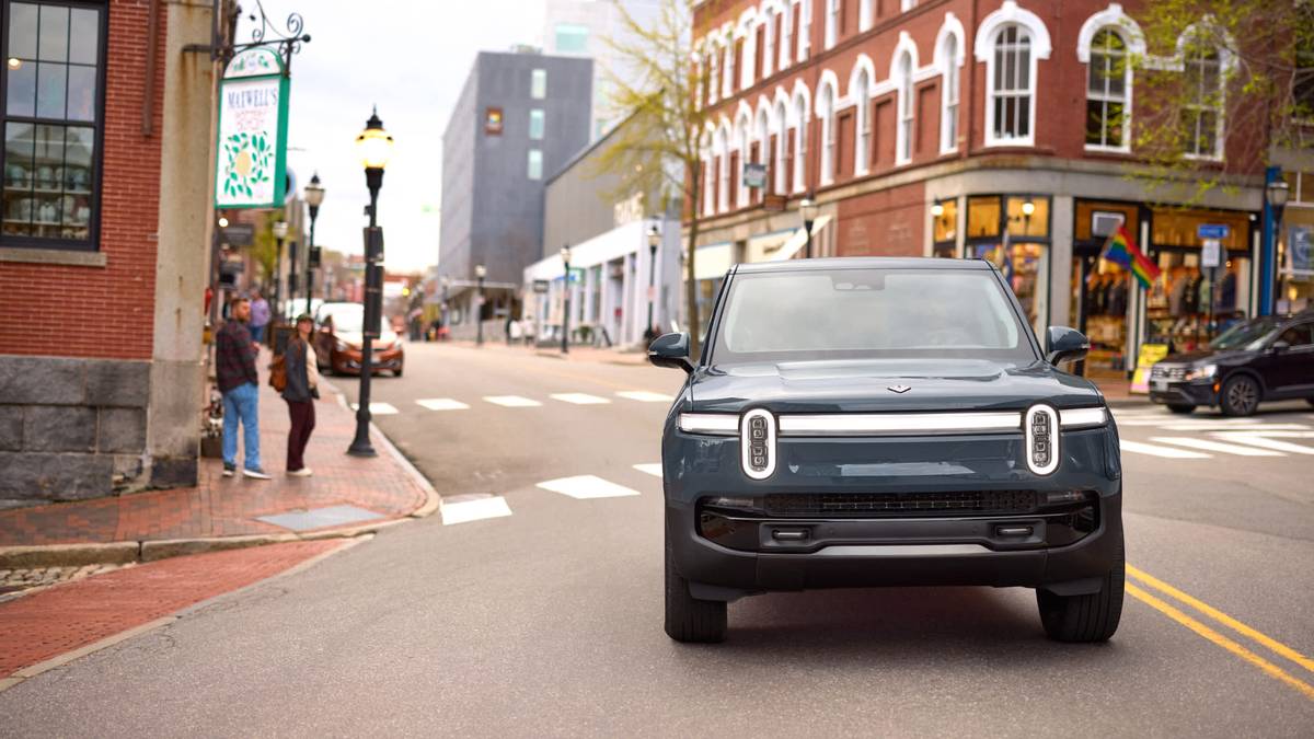 A Blue 2025 Rivian R1S driving down a city street with historic buildings, while pedestrians observe from the sidewalk.