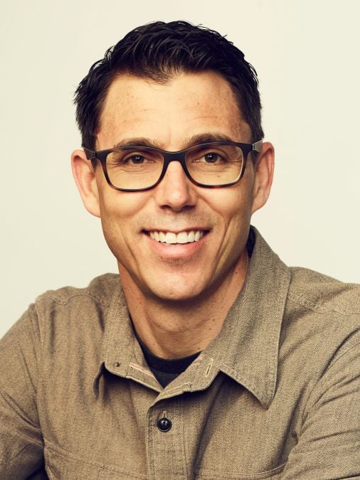 A man with dark hair, wearing glasses and a brown button-up shirt, smiles warmly at the camera. The background is plain, highlighting his friendly expression.