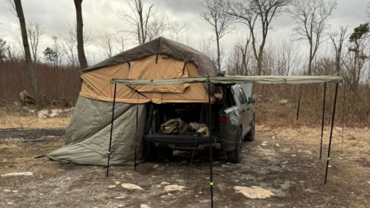 Rivian R1T with rooftop tent and side awning in bare winter forest campsite with rocky ground