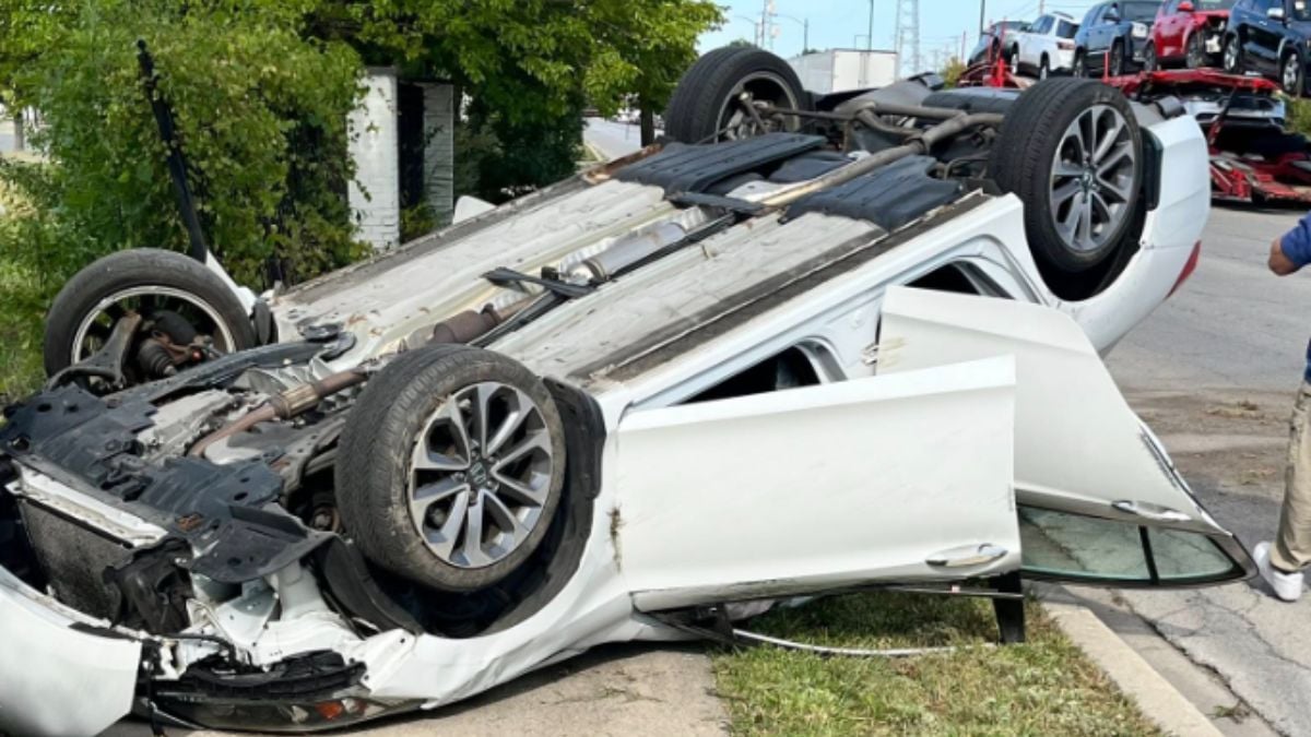 Overturned white car on a roadside, flipped upside down with wheels in the air. Nearby greenery and car carrier in background, conveying an accident scene.