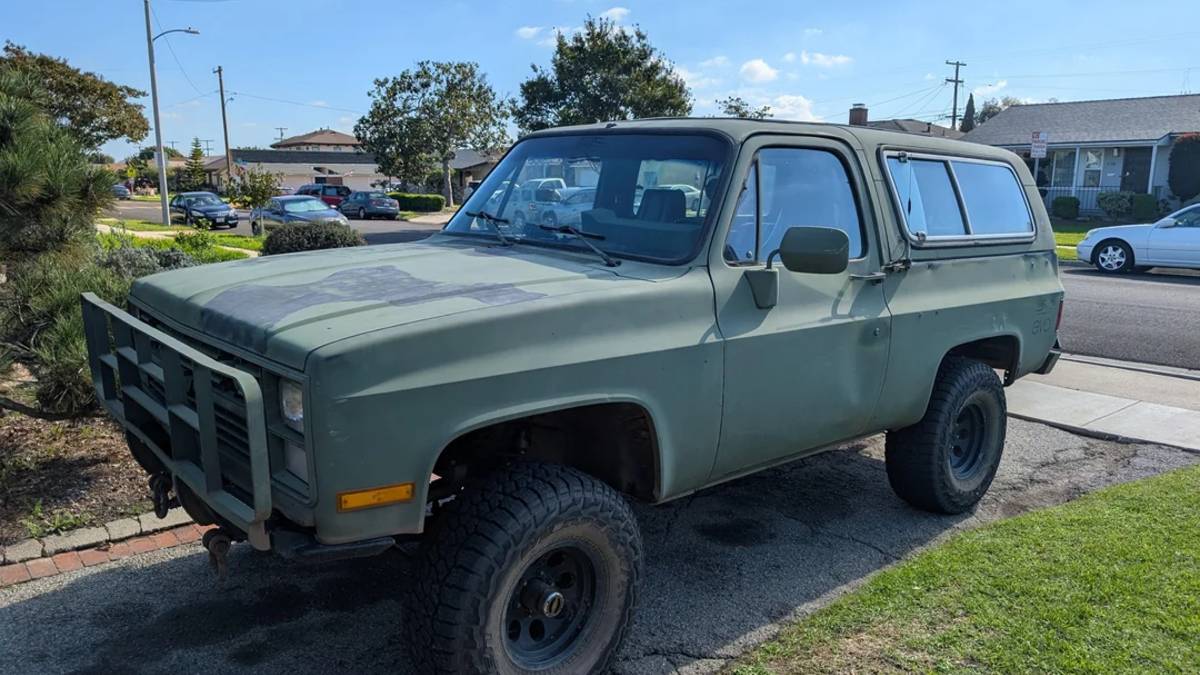 Vintage Chevrolet K5 Blazer in matte military green parked on a residential driveway, showing lifted suspension, steel wheels, and classic truck body.