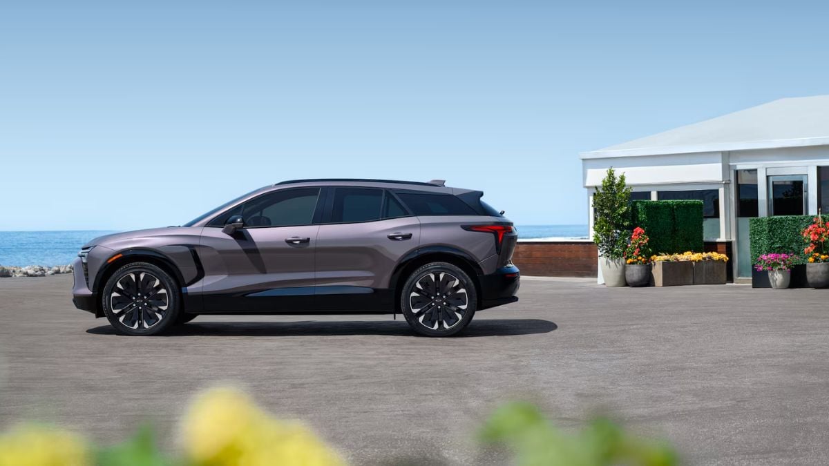 A metallic gray Chevrolet Blazer EV is shown in side profile view, parked at an oceanfront location with modern architecture and colorful landscaping in the background.