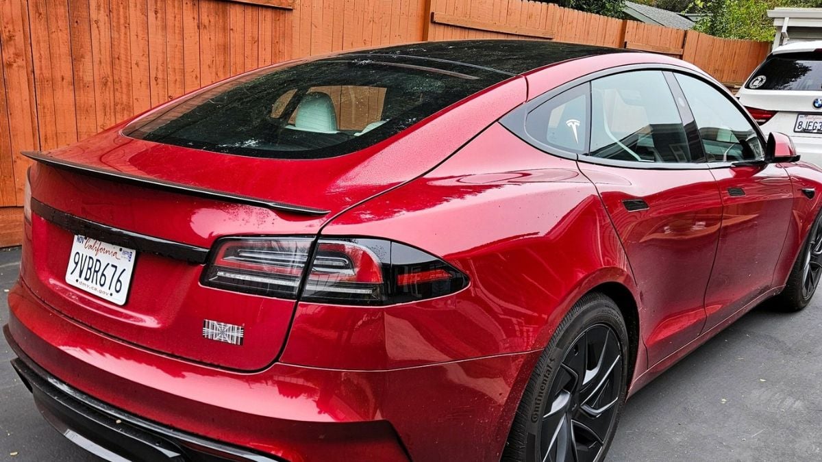 A red Tesla Model S shown from the rear three-quarter angle, featuring a black glass roof and aftermarket wheels parked next to a wooden fence.