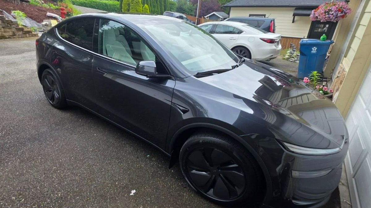 Sleek gray Tesla parked on a driveway next to a white car. Garage, recycling bin, and flowers create a suburban backdrop.