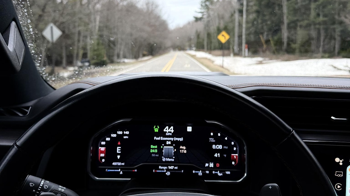 View from Dennis' GMC Sierra AT4 dashboard showing the steering wheel and digital display, focusing on fuel economy stats while driving on a calm, tree-lined road.